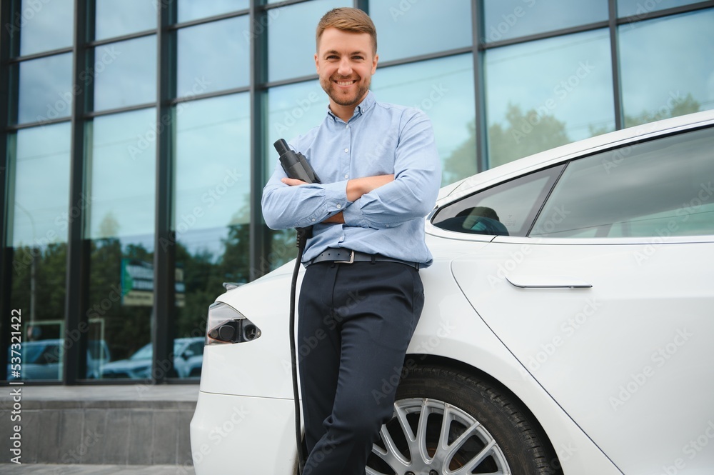 portrait of young handsome man in casual wear, standing at the charging station. Eco electric car concept.