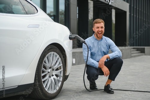 Hansome bearded guy sitting near his new modern electric car and holding plug of the charger, while car is charging at the charging station. © Serhii