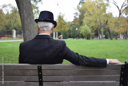 Old man with black hat sitting on a public bench in a park. Grey haired elderly man in elegant suit watching people 