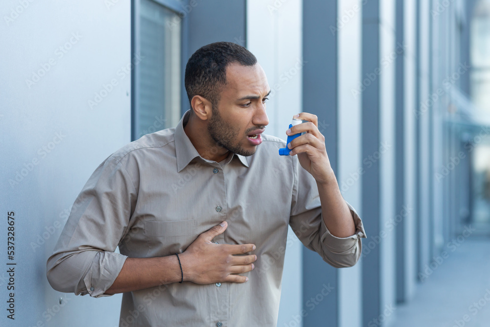 A man outside an office building has a severe asthma attack, a businessman is having difficulty breathing, a worker in a shirt is using an inhaler to make breathing easier, a hispanic man in a casual - obrazy, fototapety, plakaty 