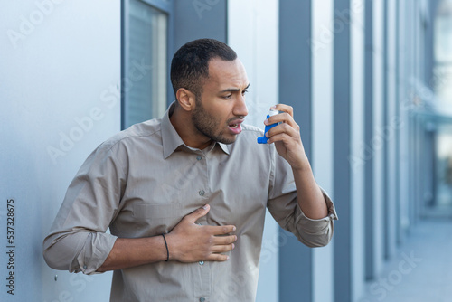 A man outside an office building has a severe asthma attack, a businessman is having difficulty breathing, a worker in a shirt is using an inhaler to make breathing easier, a hispanic man in a casual photo
