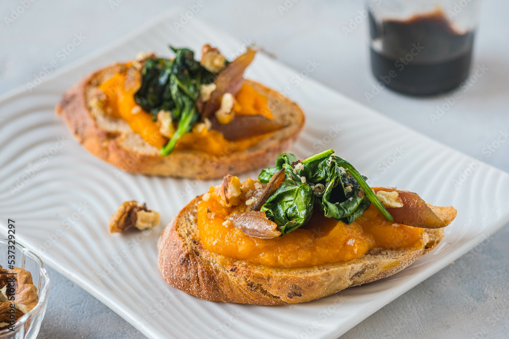 Open sandwich or bruschetta with pumpkin puree, stewed spinach and smoked fish on a white plate against a light concrete background. Antipasti.