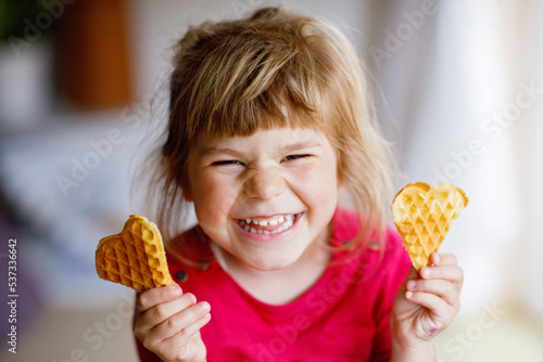 Portrait of happy little preschool girl holding fresh baked heart waffle. Smiling hungry toddler child with sweet biscuit wafer. Sweet sugar belgian waffles.