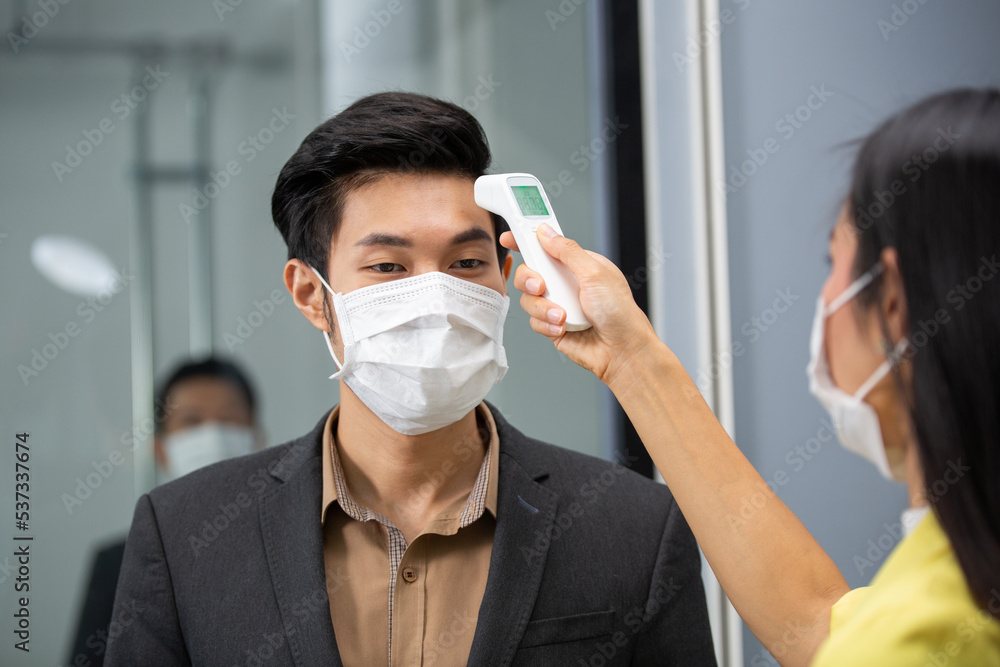 Asian employee woman using infrared thermometer check fever temperature measurement of office worker wearing face mask before enter room at entrance door. Coronavirus prevention, new normal working