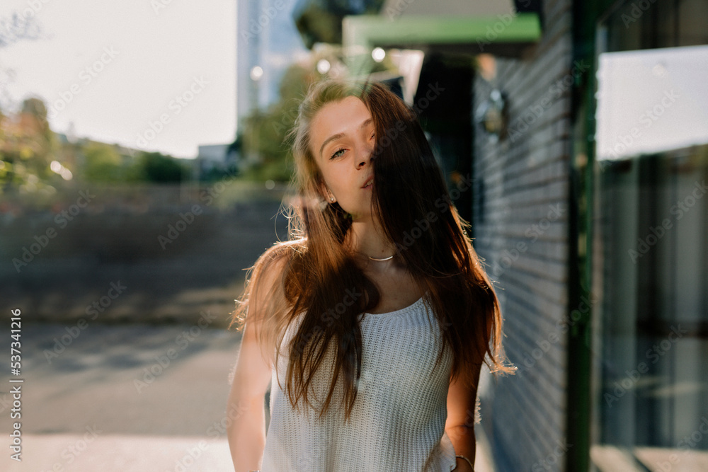 Outdoor photo of stylish young woman with loose hair wearing white t-shirt posing at camera on street inn sunny warm day. Brown-haired woman in spring day. City life concept