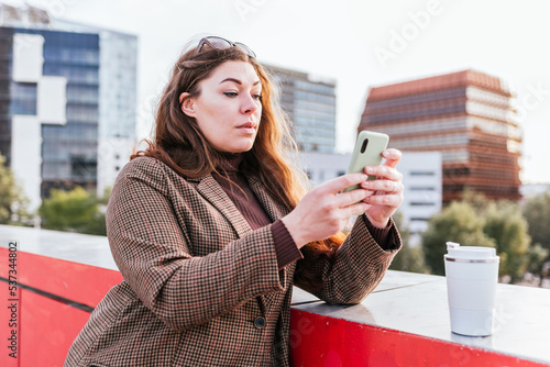 Female in outerwear with long hair leaning on border and taking picture of zero waste cup of takeaway coffee on city street in daytime