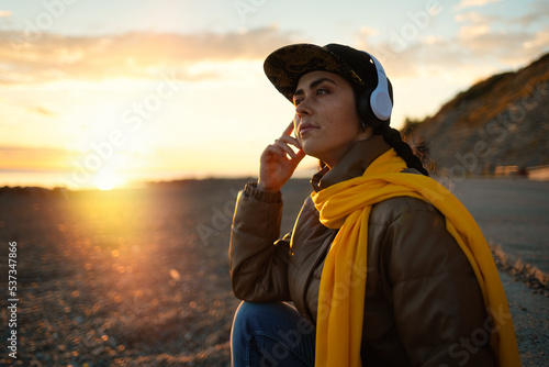Portrait of pretty young Caucasian woman in a cap listens to music with headphones. Sunset and beach in the background. The concept of freedom and psychology
