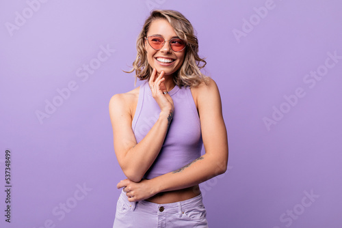 Positive young caucasian girl smiles teeth looking away posing on purple background. Blonde woman with wavy hair in sunglasses, wears top and pants. Real emotions concept