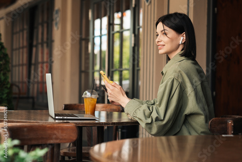 Cheerful Young Woman Holding Credit Card and Smartphone Making Online Shopping While Sitting at a Cafe Table on Terrace with Laptop and Cocktail, Looking Away.