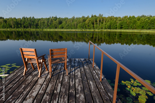 Two wooden chairs on a wooden jetty overlooking a calm lake and forest on a sunny day. The concept of relaxation,