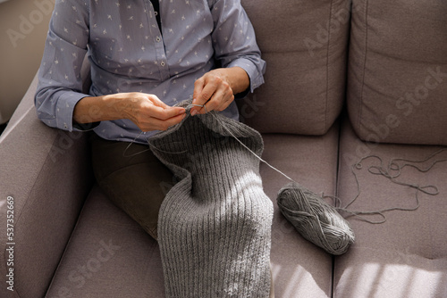 Woman knitting gray wool scarf. Needlework at home. Women’s hobby. Knitting process in women’s hands