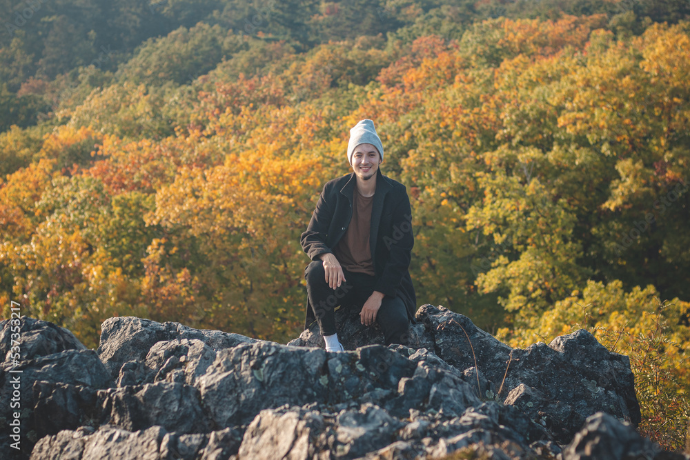 Attractive young man with smile on his mouth in a black coat and grey cap walks sits on the rock, and behind him the leafy trees play with all the colours of autumn