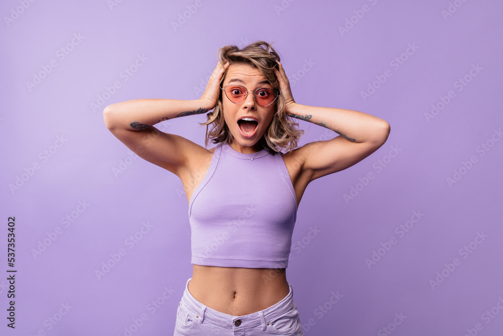 Young caucasian girl with open mouth looks surprised, holds head on purple background. Blonde wears summer crop top and sunglasses. Shock concept Young caucasian girl with open mouth looks surprised, holds head on purple background. Blonde wears summer crop top and sunglasses. Shock concept