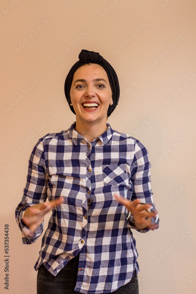 A picture of a smiling woman, wearing a black hijab and a white-blue button up t-shirt, standing in front of a beige background while explaining something A picture of a smiling woman, wearing a black hijab and a white-blue button up t-shirt, standing in front of a beige background while explaining something