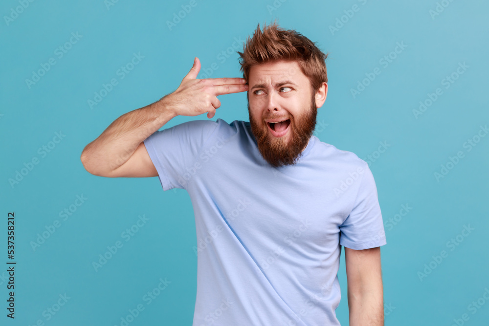 Depression, suicide gesture. Portrait of crazy bearded man standing with finger gun pointed to head, shooting himself with hand pistol. Indoor studio shot isolated on blue background. Depression, suicide gesture. Portrait of crazy bearded man standing with finger gun pointed to head, shooting himself with hand pistol. Indoor studio shot isolated on blue background.
