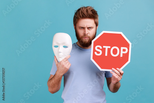 Portrait of bossy serious handsome bearded man holding red stop sign and white mask, looking at camera with strict expression. Indoor studio shot isolated on blue background. photo