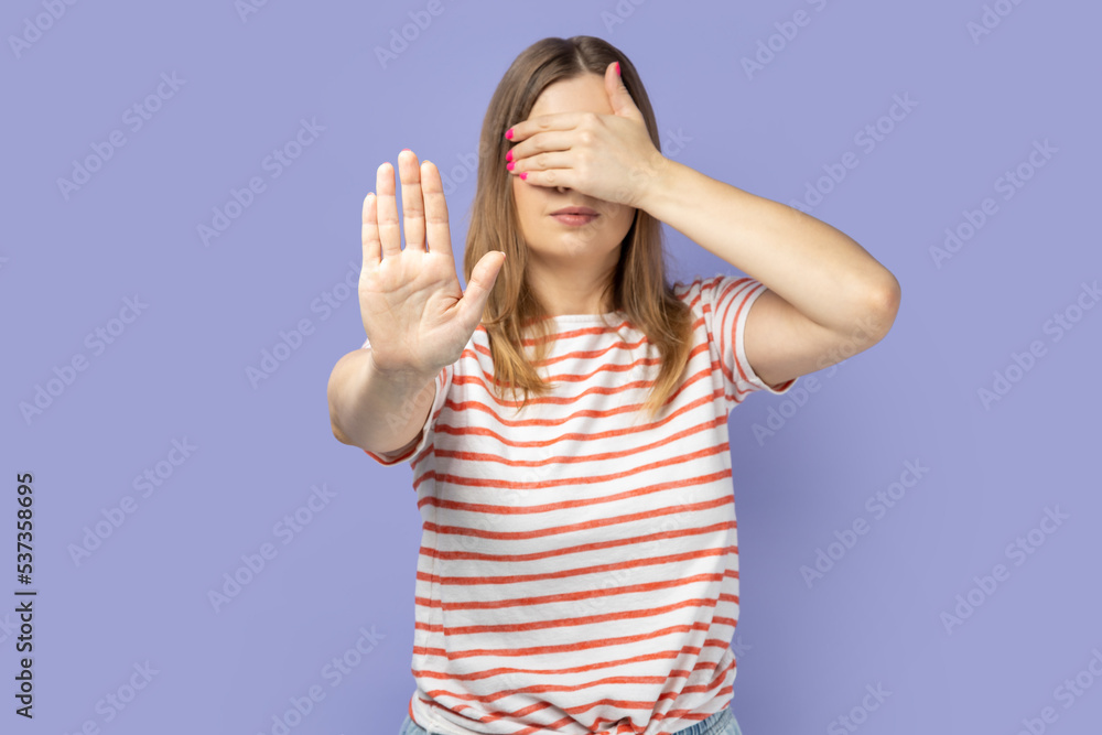 Portrait of blond woman wearing striped T-shirt showing stop gesture, covering eyes with hand, feeling shamed and scared to watch. Indoor studio shot isolated on purple background. Portrait of blond woman wearing striped T-shirt showing stop gesture, covering eyes with hand, feeling shamed and scared to watch. Indoor studio shot isolated on purple background.