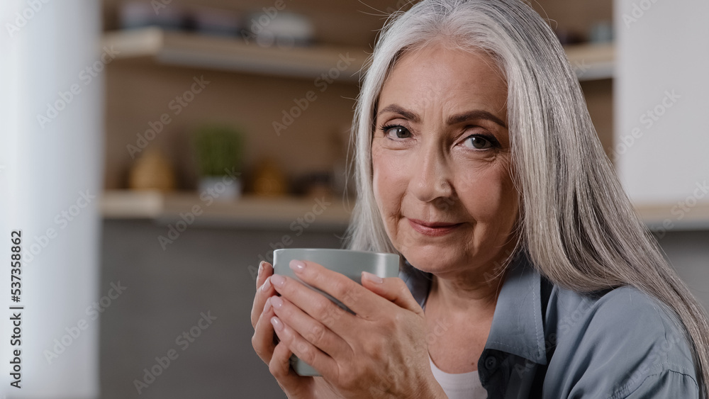 Dreamy mature woman holding cup of hot beverage warming hands on mug preventing flu looking away drinking morning coffee positive dreaming pleasant memories happy elderly grandmother relaxing at home Dreamy mature woman holding cup of hot beverage warming hands on mug preventing flu looking away drinking morning coffee positive dreaming pleasant memories happy elderly grandmother relaxing at home