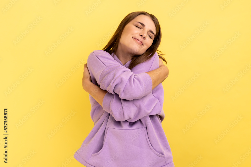 I love myself. Portrait of selfish narcissistic young woman embracing herself and smiling with dreamy satisfied expression, wearing purple hoodie. Indoor studio shot isolated on yellow background. I love myself. Portrait of selfish narcissistic young woman embracing herself and smiling with dreamy satisfied expression, wearing purple hoodie. Indoor studio shot isolated on yellow background.