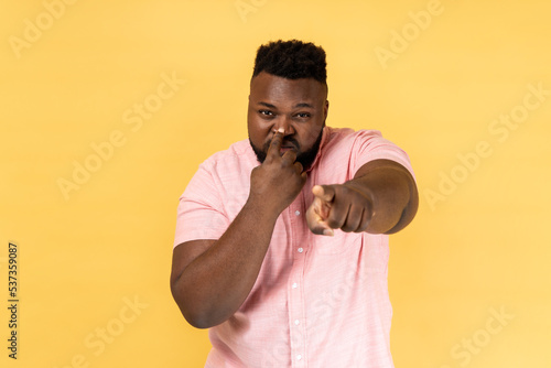 You are liar. Portrait of man wearing pink shirt standing with finger on her nose and showing lie gesture, pointing to camera. Indoor studio shot isolated on yellow background.