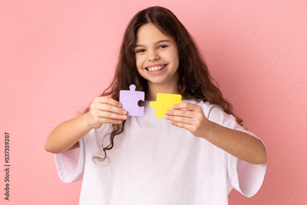 Portrait of satisfied happy smiling little girl wearing white T-shirt holding yellow and purple puzzle pieces, solving task, looking at camera. Indoor studio shot isolated on pink background. Portrait of satisfied happy smiling little girl wearing white T-shirt holding yellow and purple puzzle pieces, solving task, looking at camera. Indoor studio shot isolated on pink background.