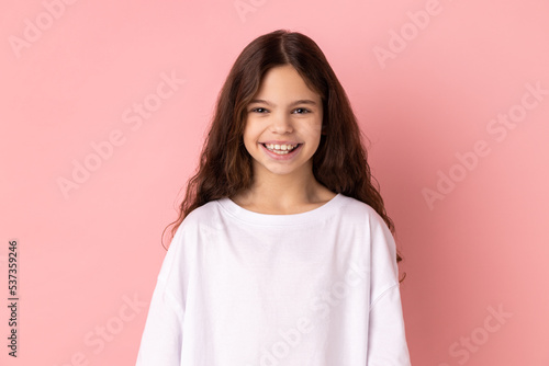 Portrait of happy satisfied little girl wearing white T-shirt looking at camera with toothy smile, expressing positive expression. Indoor studio shot isolated on pink background.
