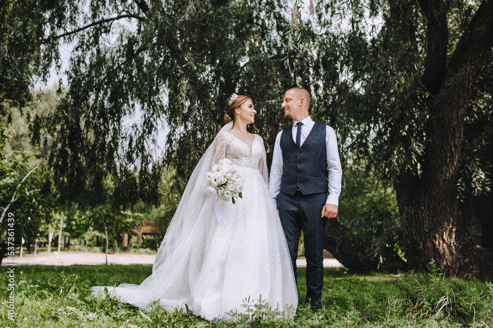 A stylish groom in a blue vest and a beautiful smiling bride are walking in a park in nature on green grass, holding hands. Wedding photo of newlyweds in love.