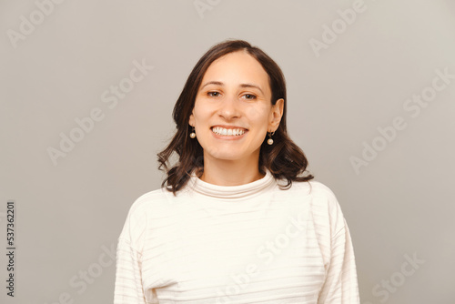 Portrait of a wide smiling confident woman wearing white sweater in a studio over light grey background.