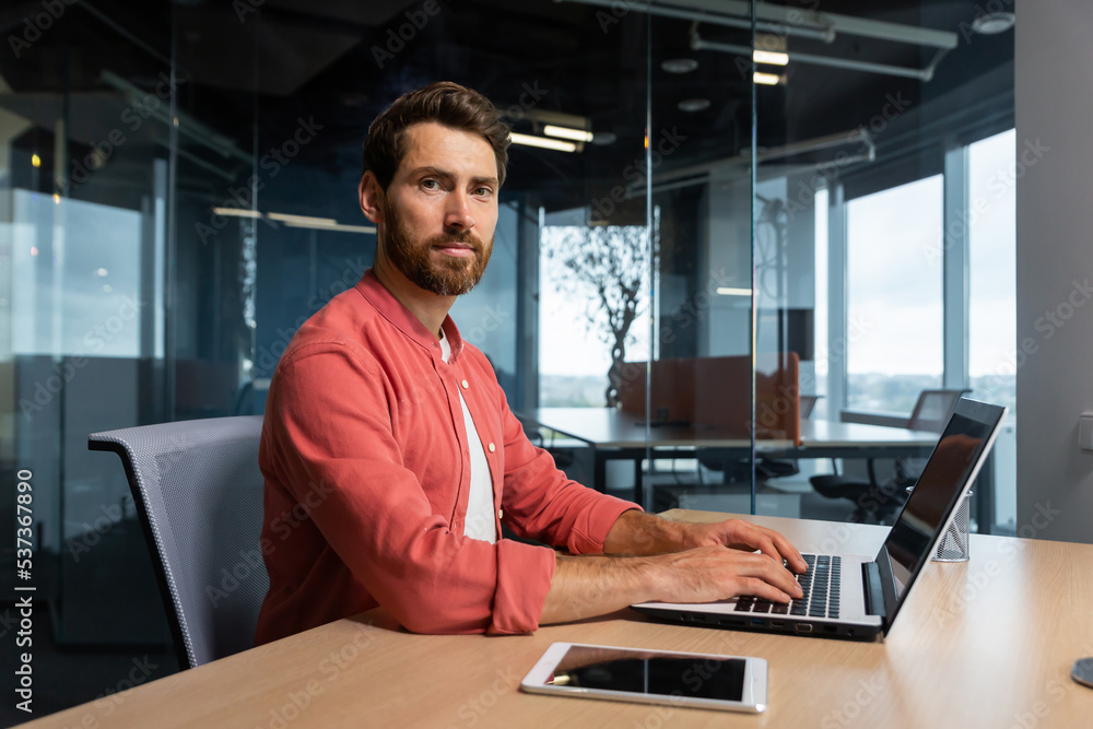 Portrait of mature programmer team leader man with beard and in red shirt is concentrating and thinking looking at camera , developer working inside modern company using laptop for coding. Portrait of mature programmer team leader man with beard and in red shirt is concentrating and thinking looking at camera , developer working inside modern company using laptop for coding.