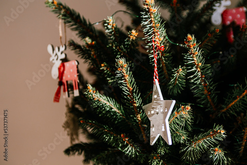Christmas tree with different wooden decorations. Christmas garland and Christmas lights. White and red colors. Christmas deer, star, snowflake. Rays of sunlight fall on the fir branches (ID: 537371475)