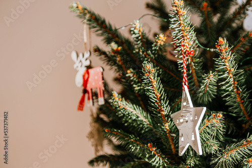 Christmas tree with different wooden decorations. Christmas garland and Christmas lights. White and red colors. Christmas deer, star, snowflake. Rays of sunlight fall on the fir branches (ID: 537371482)