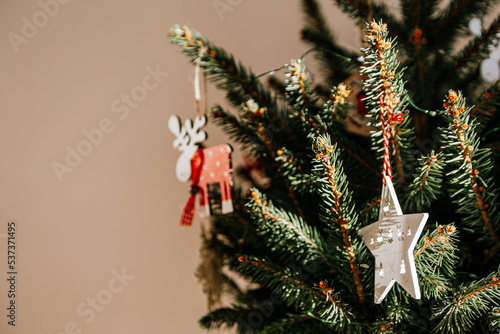 Christmas tree with different wooden decorations. Christmas garland and Christmas lights. White and red colors. Christmas deer, star, snowflake. Rays of sunlight fall on the fir branches (ID: 537371495)