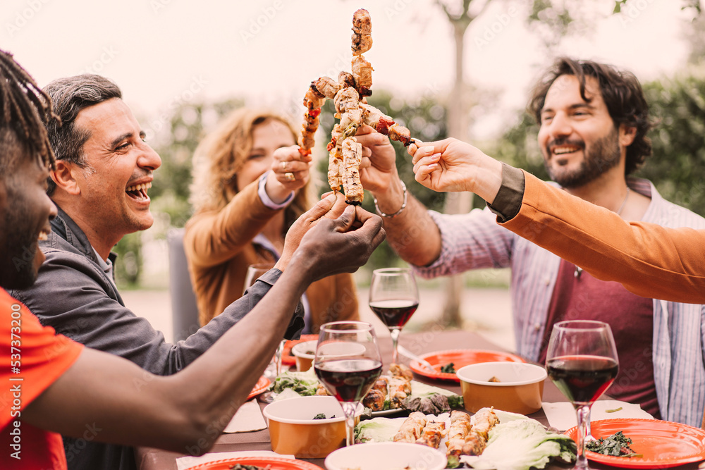Φωτογραφία Outdoor shot of multiethnic friends enjoying barbecue party in garden, sitting at the table and cheering with meat skewers
