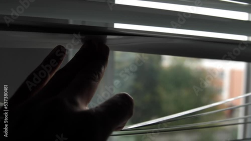 A curious man opens the roller blinds with his fingers and looks out the window. A close-up of a man's hand that opens the shutters to look at the street through the window.