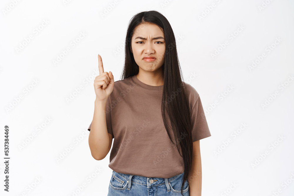 Jealous asian girl points up, grimaces from regret, disappointment, stands over white studio background Jealous asian girl points up, grimaces from regret, disappointment, stands over white studio background