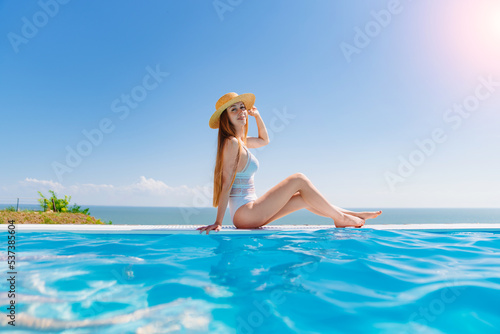 a slender woman in a white swimsuit in a straw hat sits on the pool.