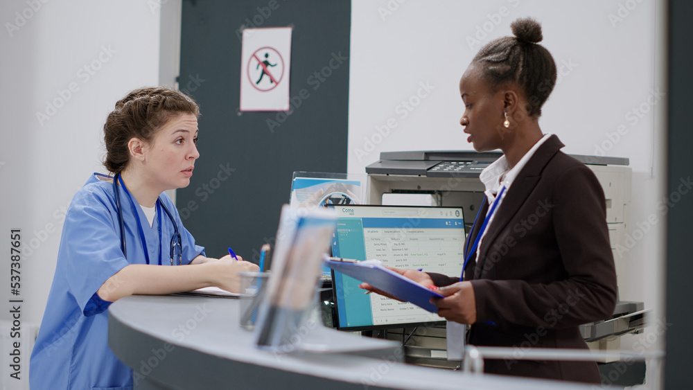 Medical assistant talking to receptionist about appointments and consultation at reception desk. Diverse hospital staff working on report papers, filling in medical forms at registration counter.