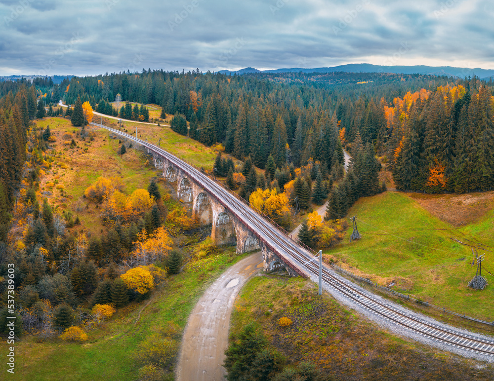 Aerial view of railway bridge, railroad, rural dirt road, green meadows, trees, hills and cloudy sky in fall. Top view of beautiful old viaduct at sunset in carpathian mountains in autumn in Ukraine