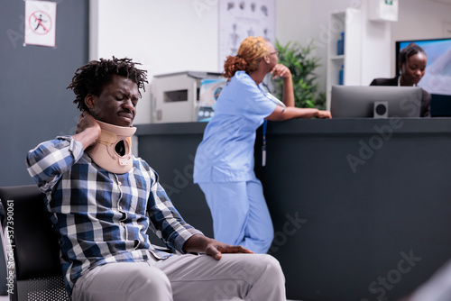 Man with cervical neck collar at reception desk, asking for help and being in pain. Patient with medical foam brace waiting to attend checkup examination appointment to cure injury after accident. photo