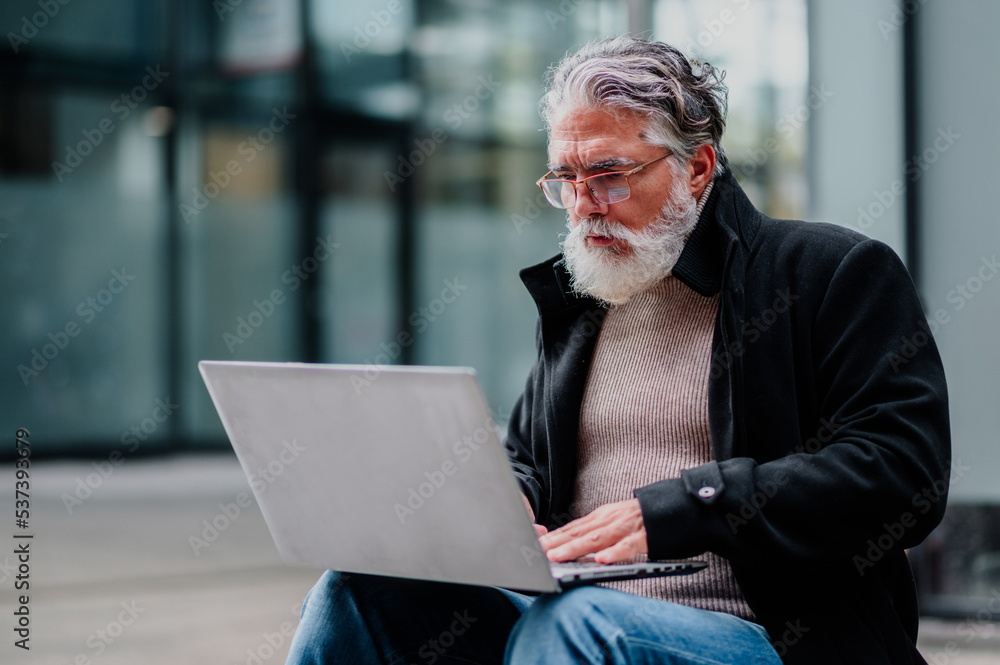 Portrait of a senior businessman using laptop and sitting on a bench outside Portrait of a senior businessman using laptop and sitting on a bench outside