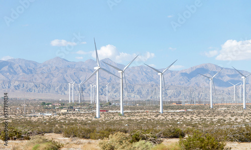 Desert landscape with windmills, low growing shrubs in the valley with mountains in background. Blue sky with clouds.