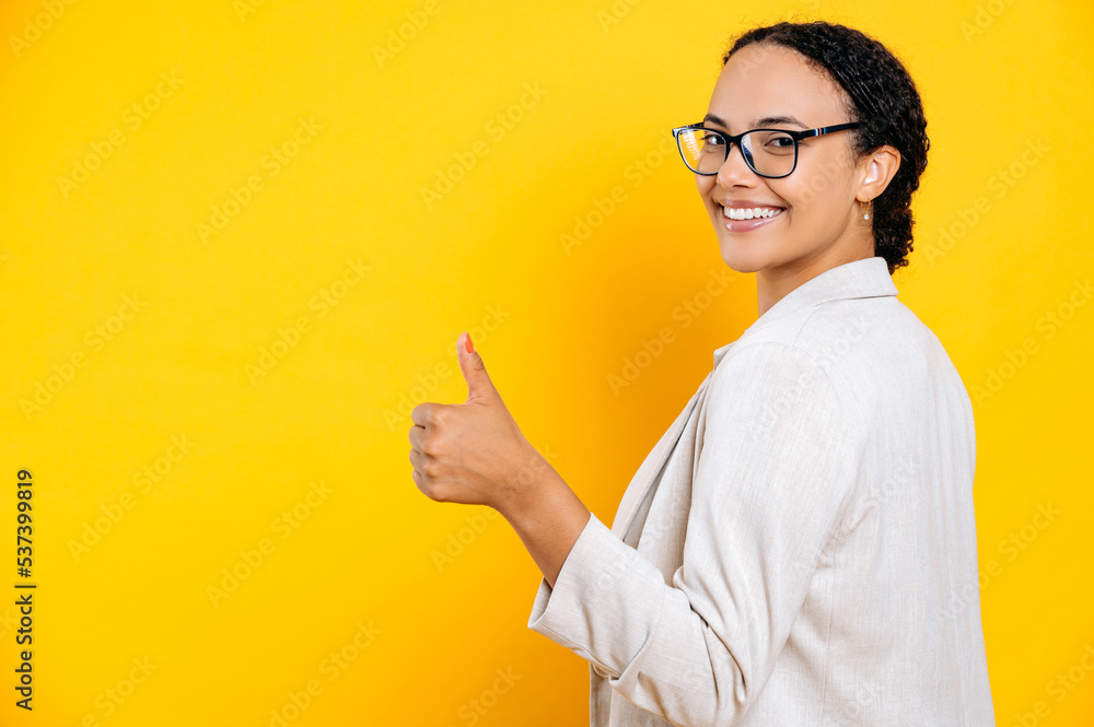 Happy successful beautiful brazilian or hispanic employee business woman, in casual jacket, stands with her back to the camera on isolated orange background, showing thumb up gesture, smiles at camera