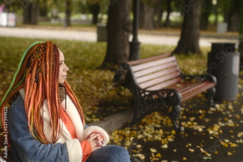 Portrait of young woman with dreadlocks sitting on bench in city park. Pretty female with colourful multicoloured hairstyle in warm clothes resting on park bench.