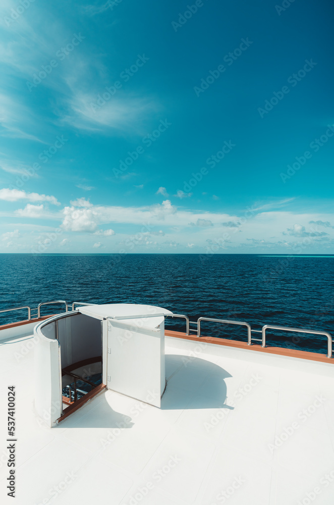 A vertical wide-angle shot of a bright diving safari yacht top deck with a selective focus on the ladder leading downstairs to the middle deck; the ocean and a blue sky in the background