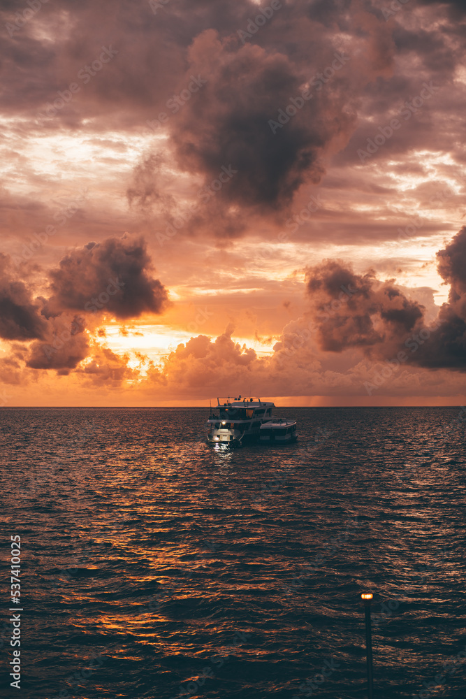 A vertical seascape with a stunning evening sky on the sunset and a silhouette of a safari diving yacht swinging on the waves aloof; a golden hour skyscape in the ocean with a boat on the water A vertical seascape with a stunning evening sky on the sunset and a silhouette of a safari diving yacht swinging on the waves aloof; a golden hour skyscape in the ocean with a boat on the water