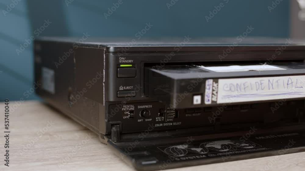 man inserts old black cassette tape into VHS player labeled CONFIDENTIAL. old vintage cassette VCR player is on table, player for viewing information on magnetic tape. man inserts old black cassette tape into VHS player labeled CONFIDENTIAL. old vintage cassette VCR player is on table, player for viewing information on magnetic tape.