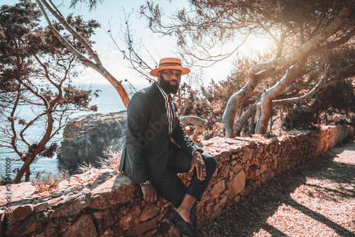 A portrait of a mature elegant bearded black man in a straw hat and a summer suit, enjoying a warm sunny day and sitting on a stone fence of a coastal park after a stroll, a shoreline in a background