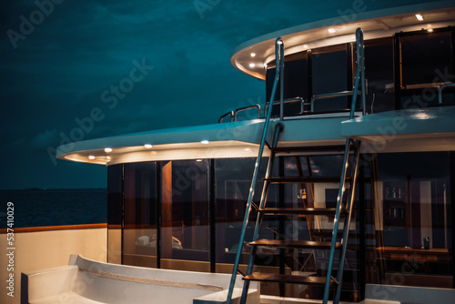 A night low-key shot of a luxury diving safari yacht deck lit by multiple ceiling lamps, with a ladder in front and a glass row of windows with a wardroom behind it; a twilight sky in the background photo