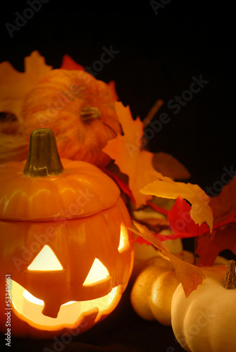  Halloween still life with ceramic jack-o-lantern with a glowing candle and artificial guords and autumn leaves all on a black background