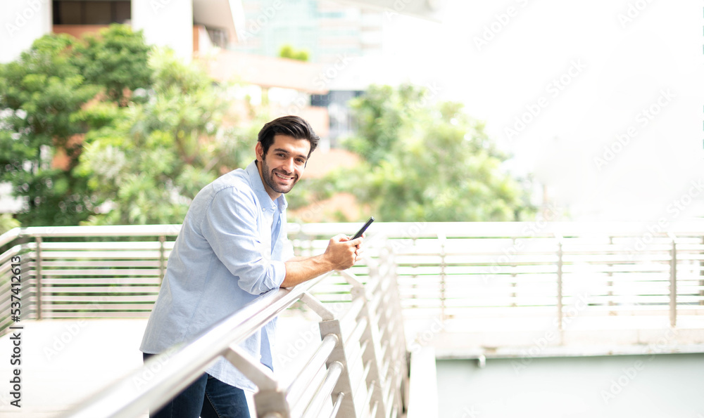 The young man holding a smartphone to waiting somethig. The man wearing casual cloth at the outside. The young man holding a smartphone to waiting somethig. The man wearing casual cloth at the outside.
